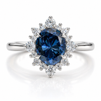 Round blue sapphire halo engagement ring with silver band, featuring a deep blue center gemstone surrounded by sparkling diamond accents in a floral cluster setting.