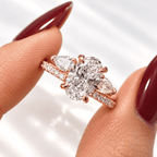 Close-up of fingers holding a rose gold ring with an oval center diamond, two pear-shaped diamonds, and a pavé diamond band, with dark red nails