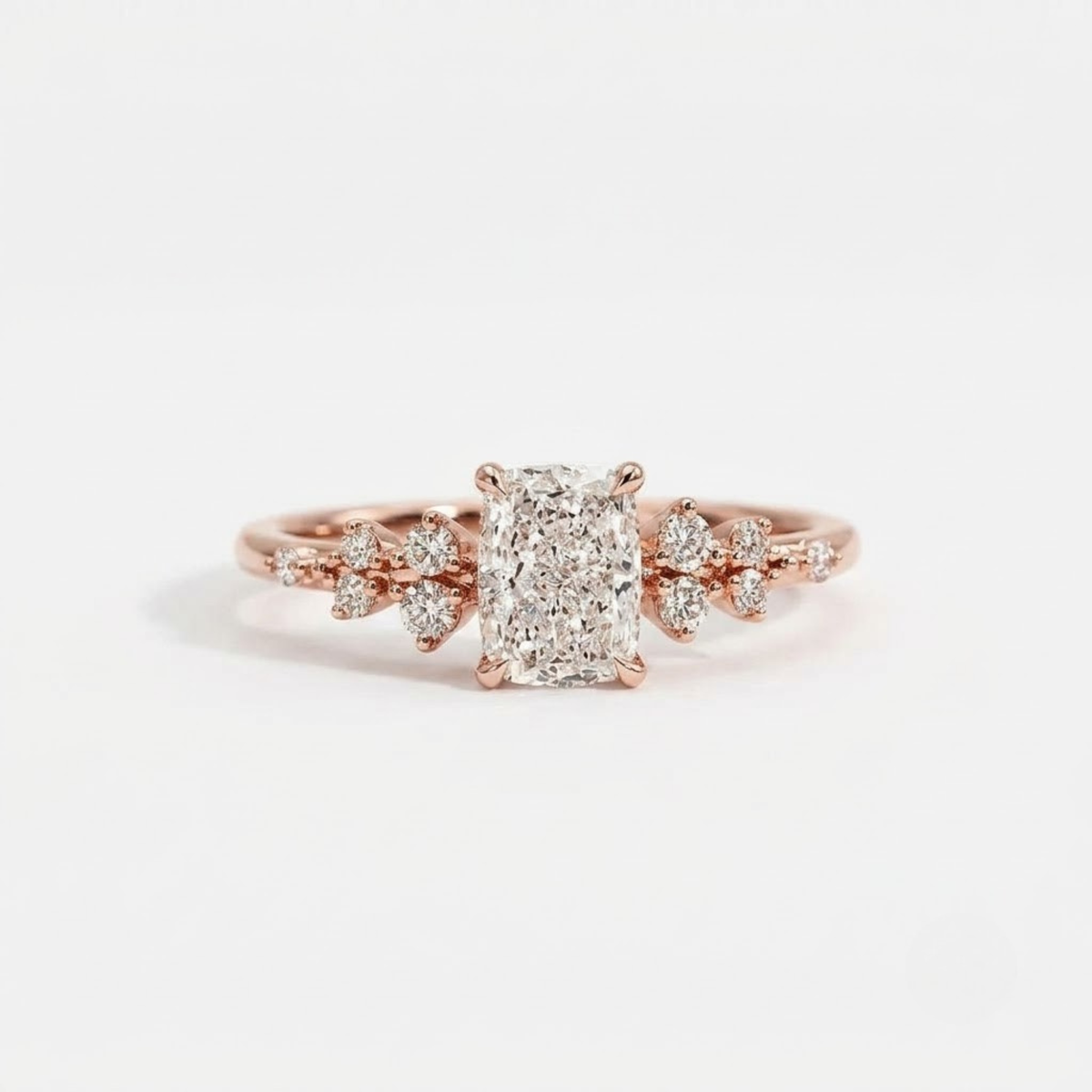 A high-resolution product photograph of a delicate yellow gold engagement ring, featuring a central cushion-cut diamond flanked by unique, clustered diamond clusters in a floral-inspired design on both shoulders. The ring is lying on a polished, reflective white surface with a soft, clean white background.