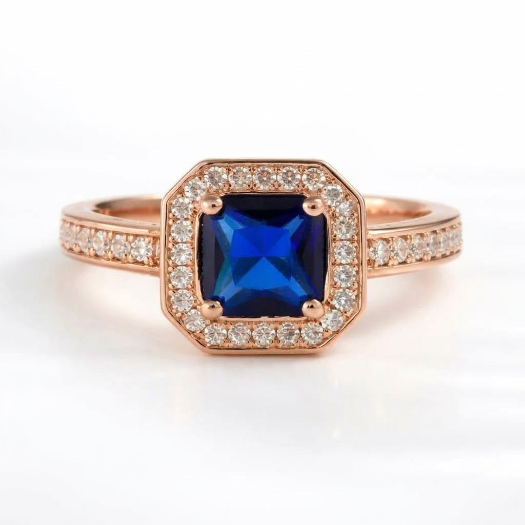 A rose gold ring featuring a square-cut blue sapphire center stone surrounded by a halo of small clear diamonds, with a diamond-accented band, shown on a white background.