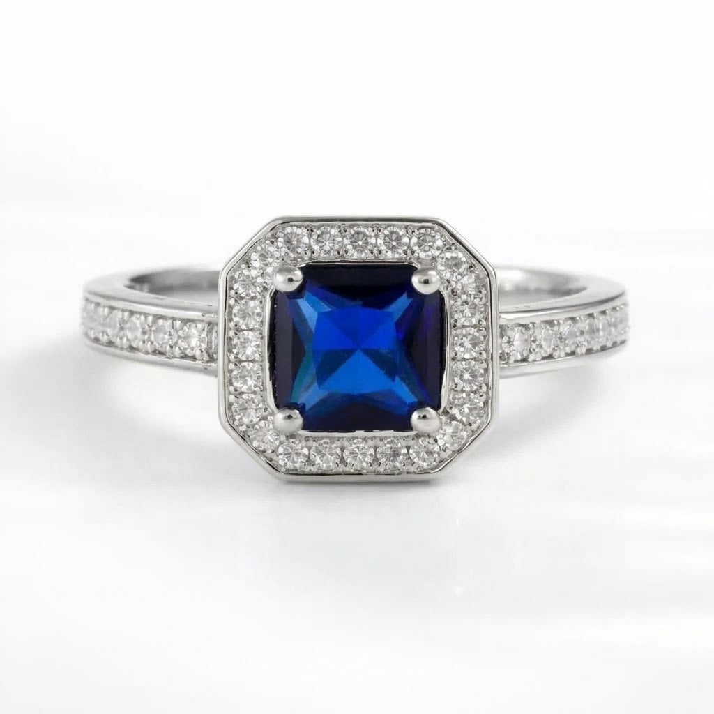 An octagonal halo ring featuring a deep blue cushion-cut center stone surrounded by a halo of small clear stones, with a silver-colored band paved with additional clear stones, set against a plain white background.