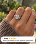 Hand with light pink nails wearing a white gold oval diamond solitaire engagement ring stacked with a slim wedding band set with small round diamonds, shown in a brown knit sleeve in front of green foliage, with a four-star customer review from Ruby overlaid at the bottom