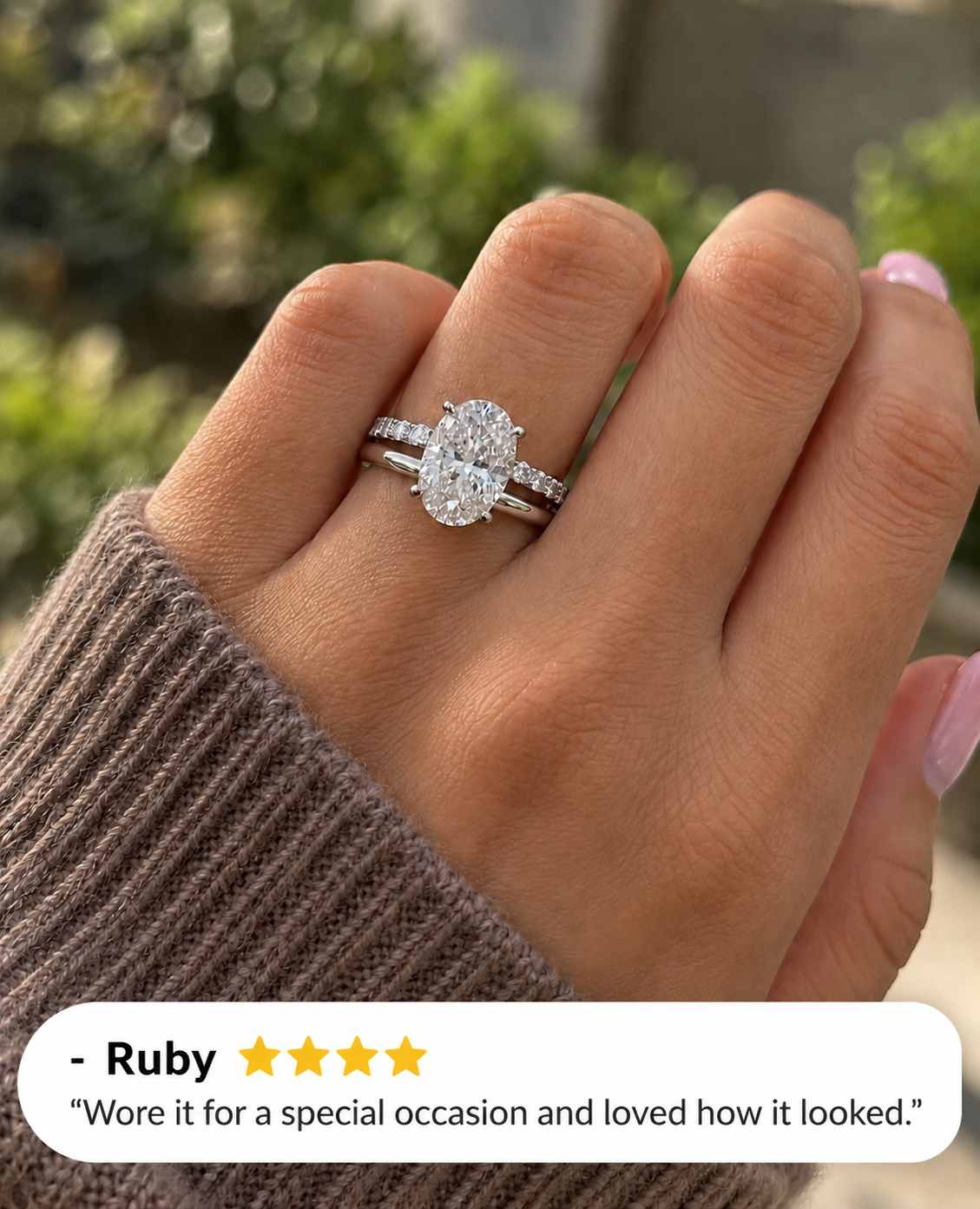 Hand with light pink nails wearing a white gold oval diamond solitaire engagement ring stacked with a slim wedding band set with small round diamonds, shown in a brown knit sleeve in front of green foliage, with a four-star customer review from Ruby overlaid at the bottom