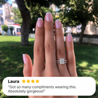 Hand with a French manicure wearing a white gold radiant cut diamond engagement ring with baguette side stones, stacked with a slim pavé diamond wedding band, held up in front of green trees in a park, with a four-star customer review from Laura overlaid at the bottom