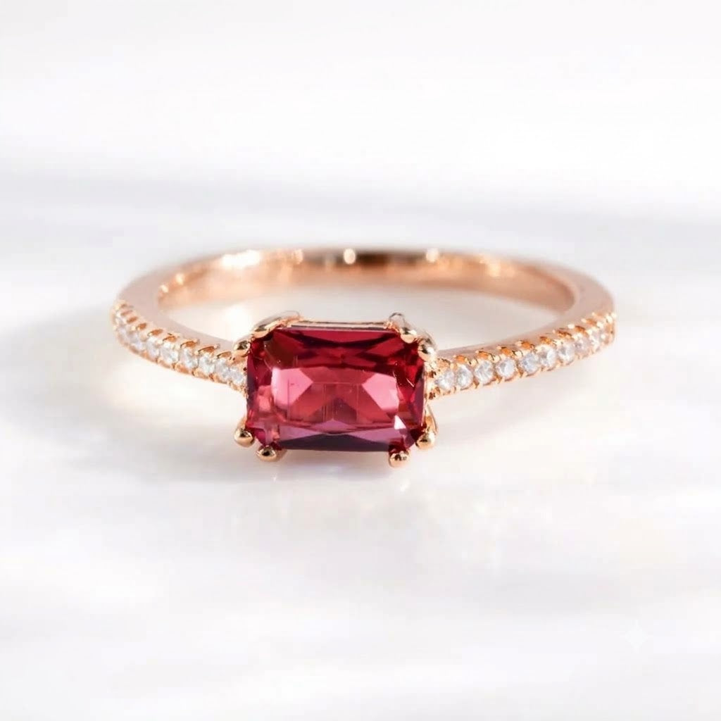 A rose gold ring featuring a rectangular emerald-cut red gemstone in a prong setting, with a pavé band of small clear stones on either side, shown on a white background.