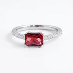 A silver ring featuring a rectangular emerald-cut red gemstone in the center, with small clear stones set along the shoulders, shown on a plain white background.