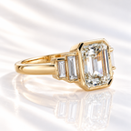 Angled view of an emerald cut diamond center stone in a yellow gold bezel setting with graduated baguette side stones along the shoulders