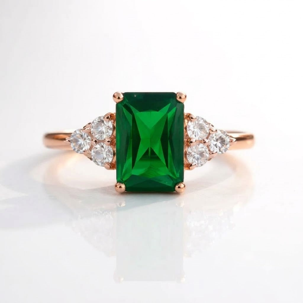 A rose gold ring featuring a rectangular emerald-cut green gemstone in the center, flanked by small clear round-cut stones on a white reflective background.