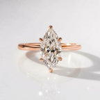 A close-up product photograph of a delicate engagement ring in 18K rose gold vermeil, featuring a large oval solitaire diamond held by four prongs, resting on a white reflective surface with soft lighting.