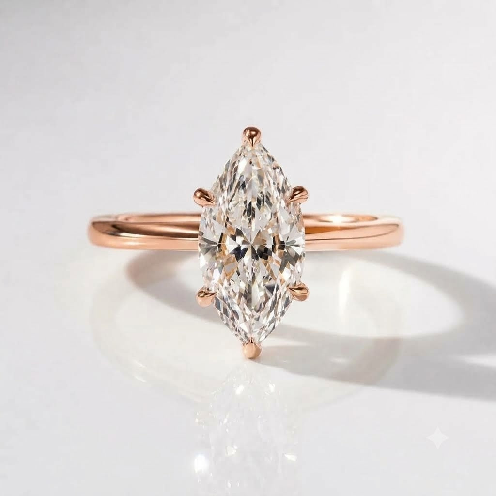A close-up product photograph of a delicate engagement ring in 18K rose gold vermeil, featuring a large oval solitaire diamond held by four prongs, resting on a white reflective surface with soft lighting.