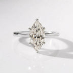 A close-up product photograph of a delicate engagement ring in polished silver, featuring a large oval solitaire diamond held by four prongs, resting on a white reflective surface with soft lighting.