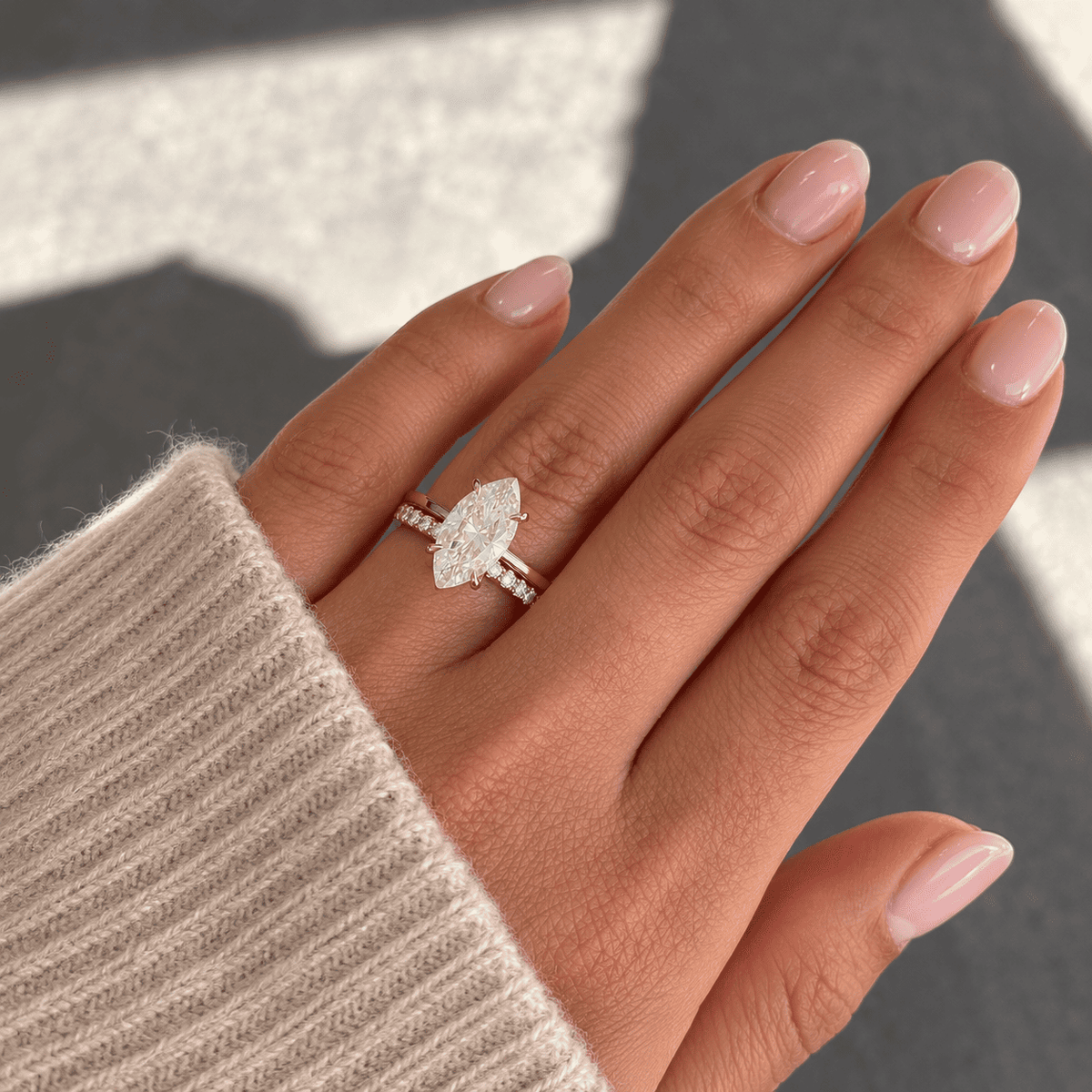 Hand with a soft pink manicure wearing a rose gold marquise diamond engagement ring stacked with a diamond wedding band, beside a cream knit sleeve.