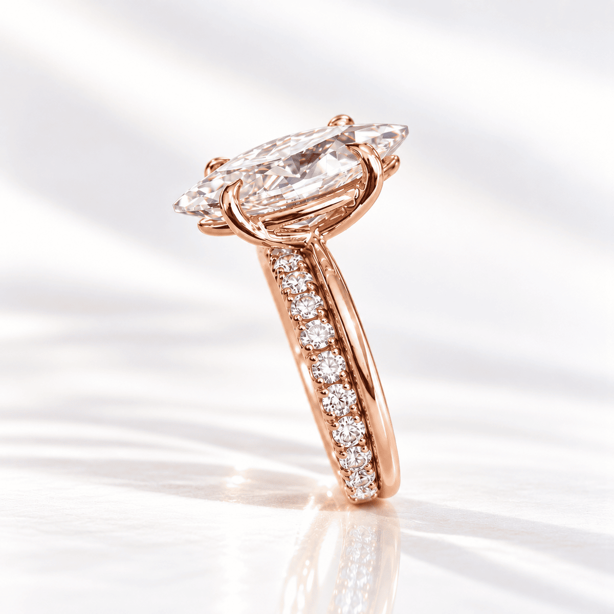 Side profile of a marquise diamond engagement ring with a pavé diamond band, set in rose gold on a glossy white surface.