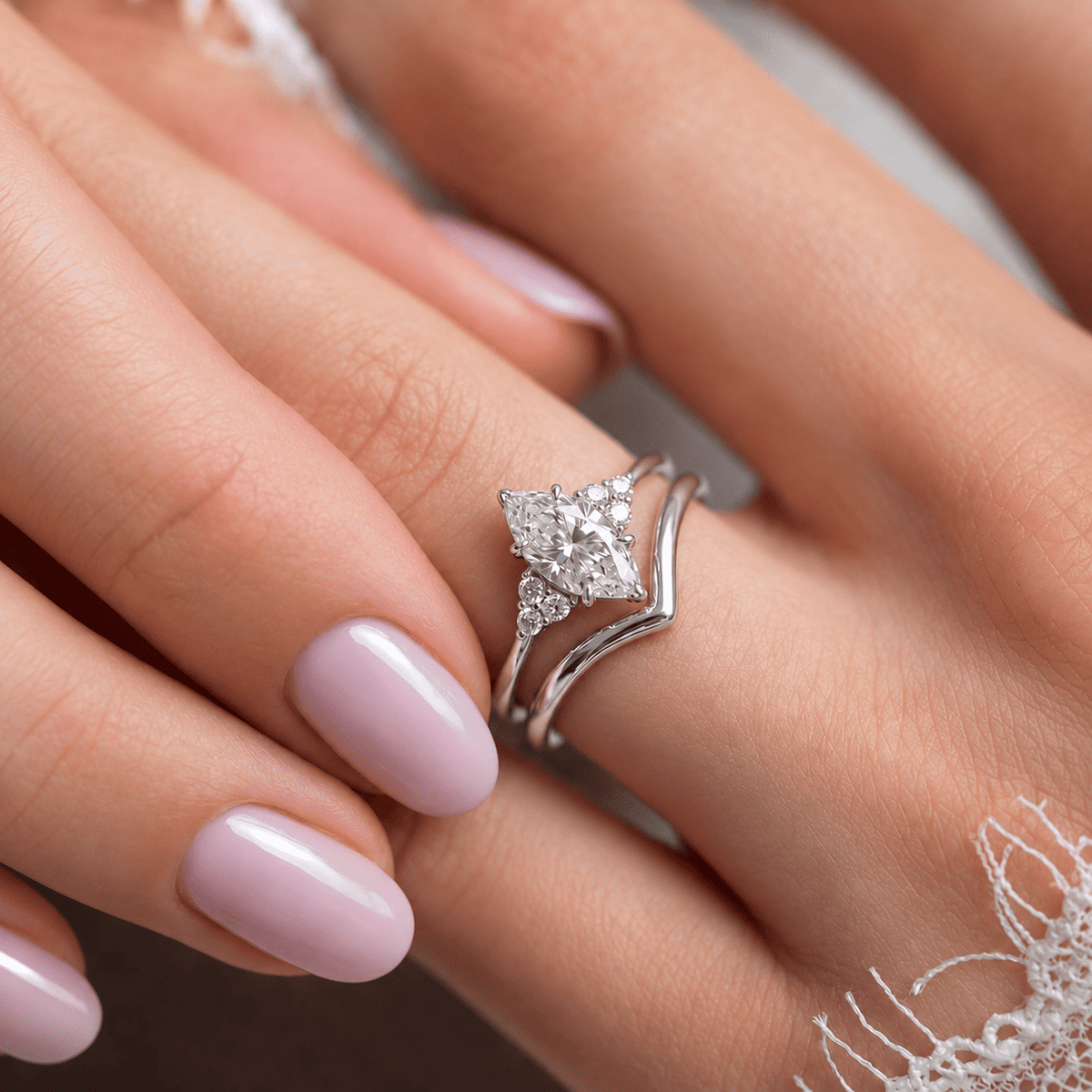 Close-up of a hand wearing a white gold marquise diamond ring stacked with a V-shaped chevron wedding band, with light pink manicured nails and white lace in the background.