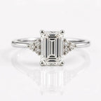 An emerald-cut solitaire diamond ring set in rose gold with two tri-stone clusters of round diamonds on either side of the center stone, resting on a white, reflective surface.