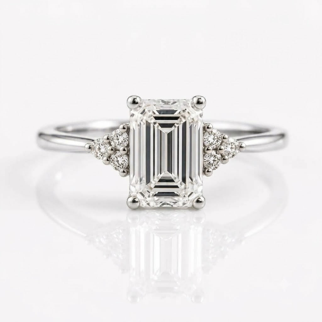 An emerald-cut solitaire diamond ring set in rose gold with two tri-stone clusters of round diamonds on either side of the center stone, resting on a white, reflective surface.
