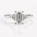 An emerald-cut solitaire diamond ring set in rose gold with two tri-stone clusters of round diamonds on either side of the center stone, resting on a white, reflective surface.