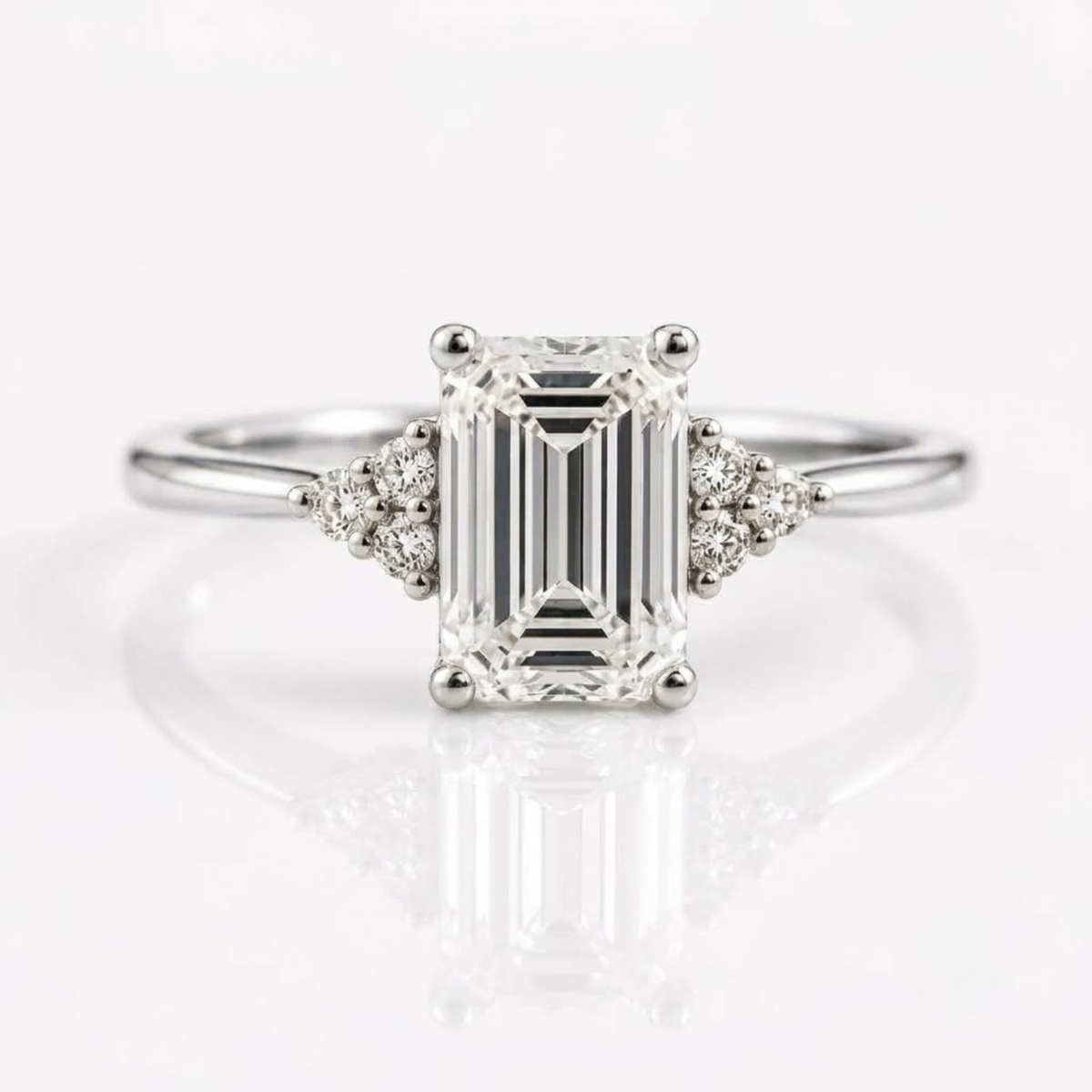 An emerald-cut solitaire diamond ring set in rose gold with two tri-stone clusters of round diamonds on either side of the center stone, resting on a white, reflective surface.