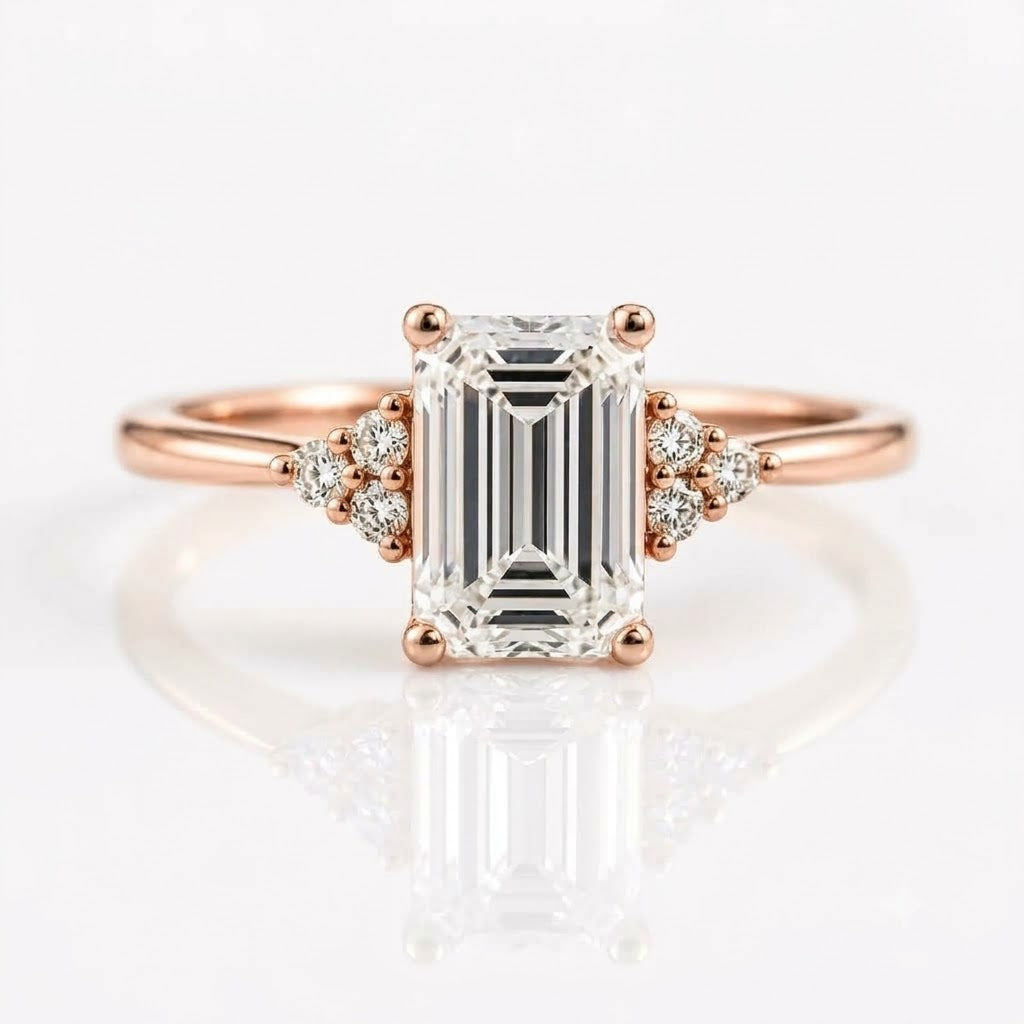 An emerald-cut solitaire diamond ring set in rose gold with two tri-stone clusters of round diamonds on either side of the center stone, resting on a white, reflective surface.