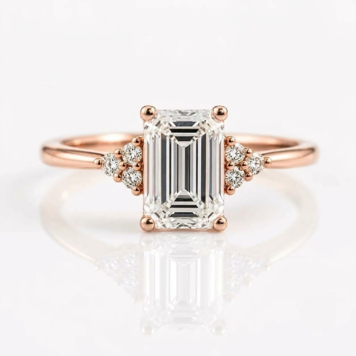 An emerald-cut solitaire diamond ring set in rose gold with two tri-stone clusters of round diamonds on either side of the center stone, resting on a white, reflective surface.