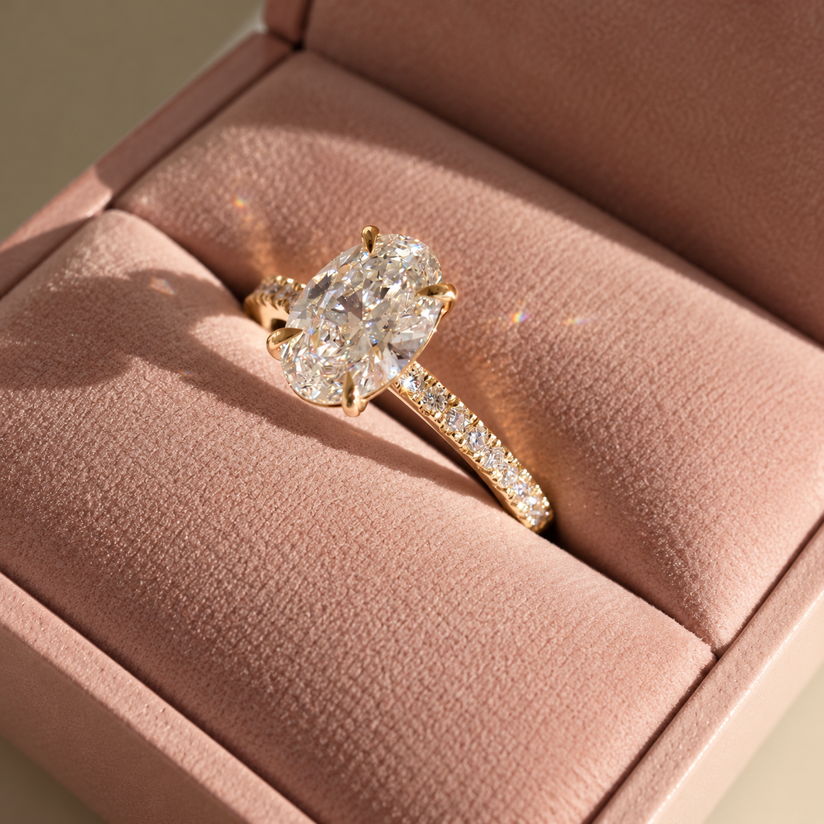Angled view of an oval cut diamond center stone in a four-prong yellow gold setting with a pavé diamond band, resting in a pink ring box