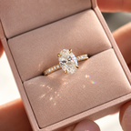Oval cut diamond center stone in a four-prong yellow gold setting with a pavé diamond band, displayed in a pink ring box