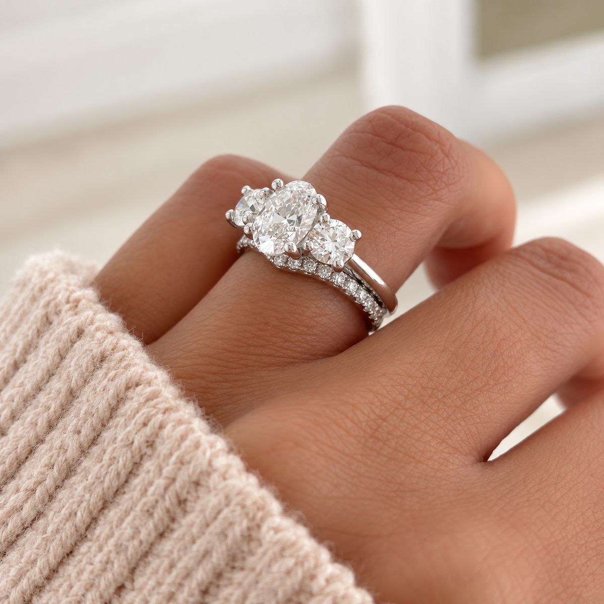 Oval cut diamond center stone in a prong white gold setting, flanked by a round side stone on each side, stacked with a pavé diamond band