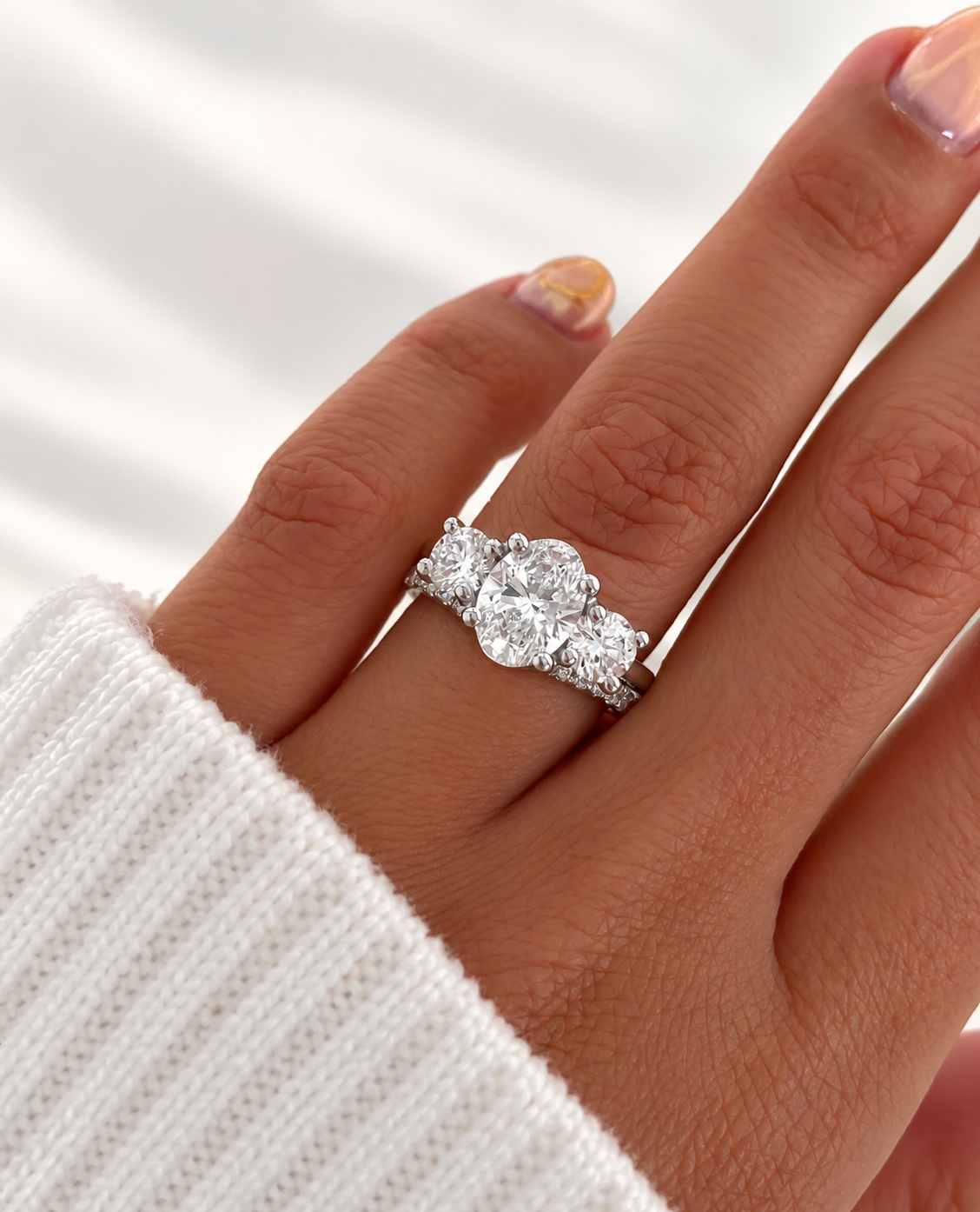 Oval cut diamond center stone in a prong white gold setting, flanked by a round side stone on each side, stacked with a pavé diamond band