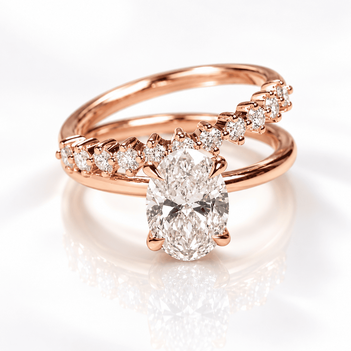 Rose gold bypass ring set with an oval center diamond and a sweeping row of graduated round diamonds, photographed on a soft white surface with a mirrored reflection.