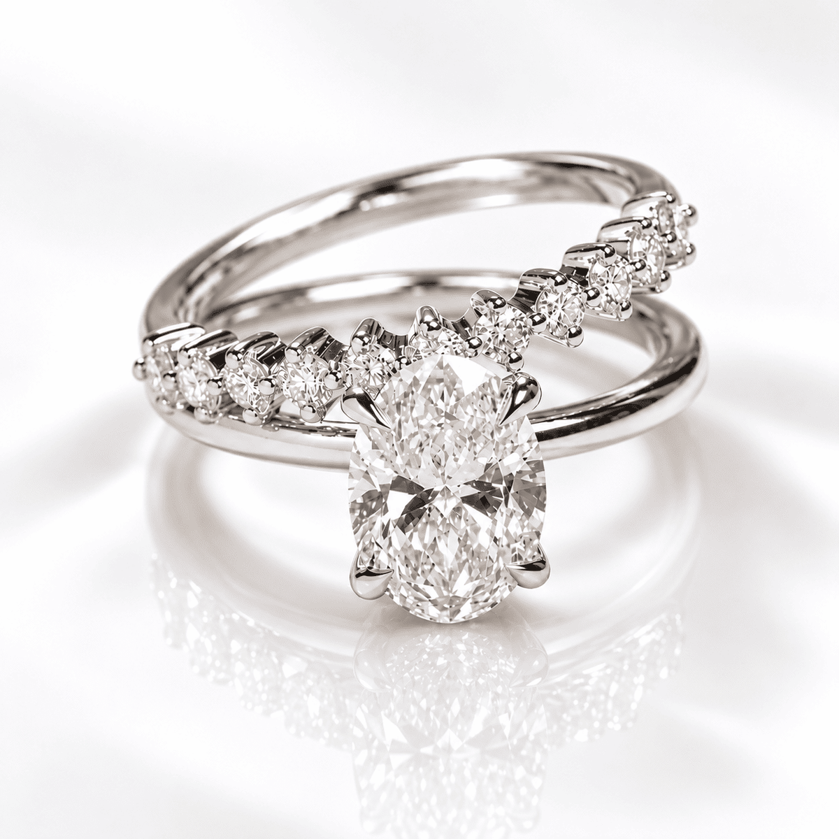 White gold or platinum bypass engagement ring featuring an oval diamond solitaire and a curved band of graduated prong-set round diamonds, displayed on a glossy white background.