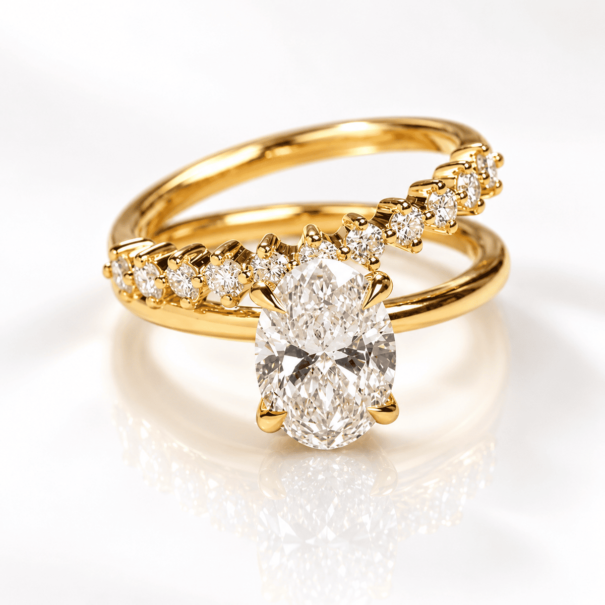 Yellow gold bypass engagement ring with a center oval diamond in a four-prong setting, paired with a curved band of graduated round diamonds, shown on a white reflective surface.