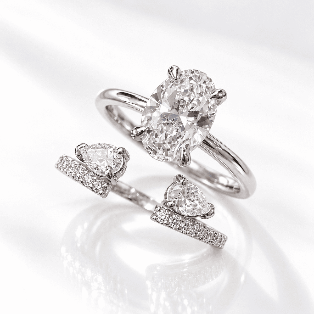 White gold open bypass ring with a four-prong oval diamond solitaire, two pear-shaped diamonds, and pavé diamond band ends, shown on a white glossy surface.