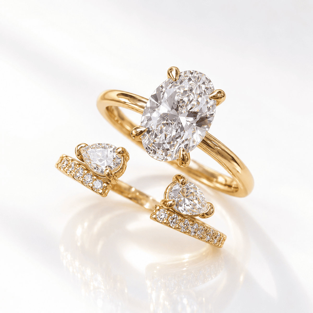 Yellow gold open bypass ring with a four-prong oval diamond solitaire, two pear-shaped diamonds, and pavé diamond band ends, shown on a white glossy surface