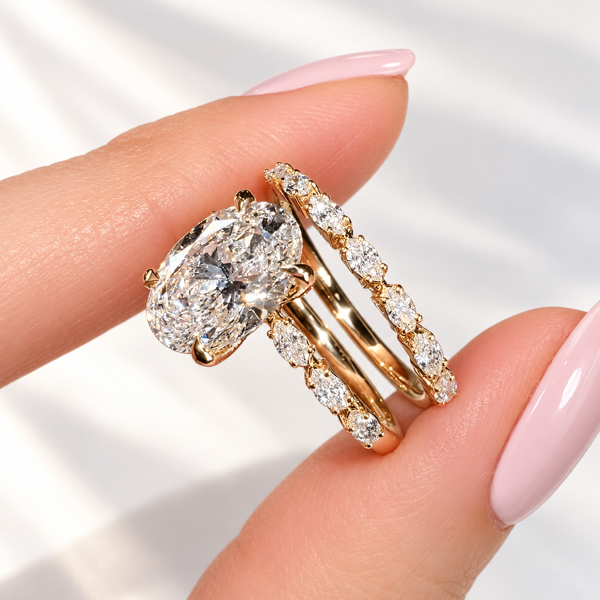 Side view of a yellow gold bridal set held between two fingers, with an oval diamond ring with marquise side stones beside a marquise diamond wedding band