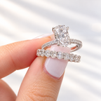 Pink-manicured fingers holding a white gold oval diamond engagement ring with a pavé band, stacked with a wedding band of oval diamonds
