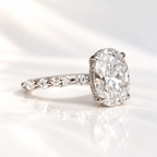White gold oval diamond engagement ring with four prongs and a hidden halo, with marquise and round diamonds along the band, resting on a glossy white surface