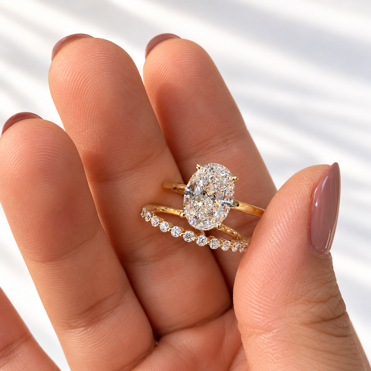 Open hand with mauve nails holding a yellow gold oval diamond solitaire engagement ring beside a slim wedding band set with round diamonds