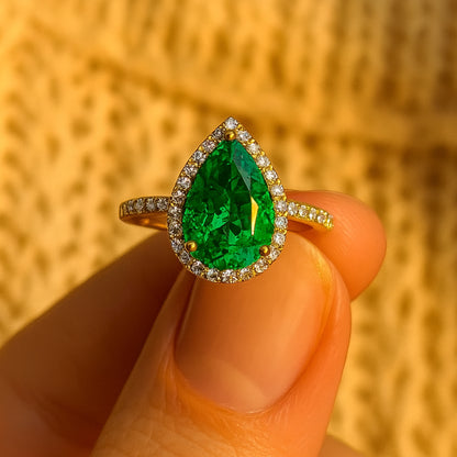 Pear-shaped green emerald ring with diamond halo and gold band held between fingers.”
