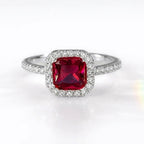 A silver-toned halo ring featuring a cushion-cut red gemstone center stone surrounded by a halo of small clear stones, set against a clean white background.