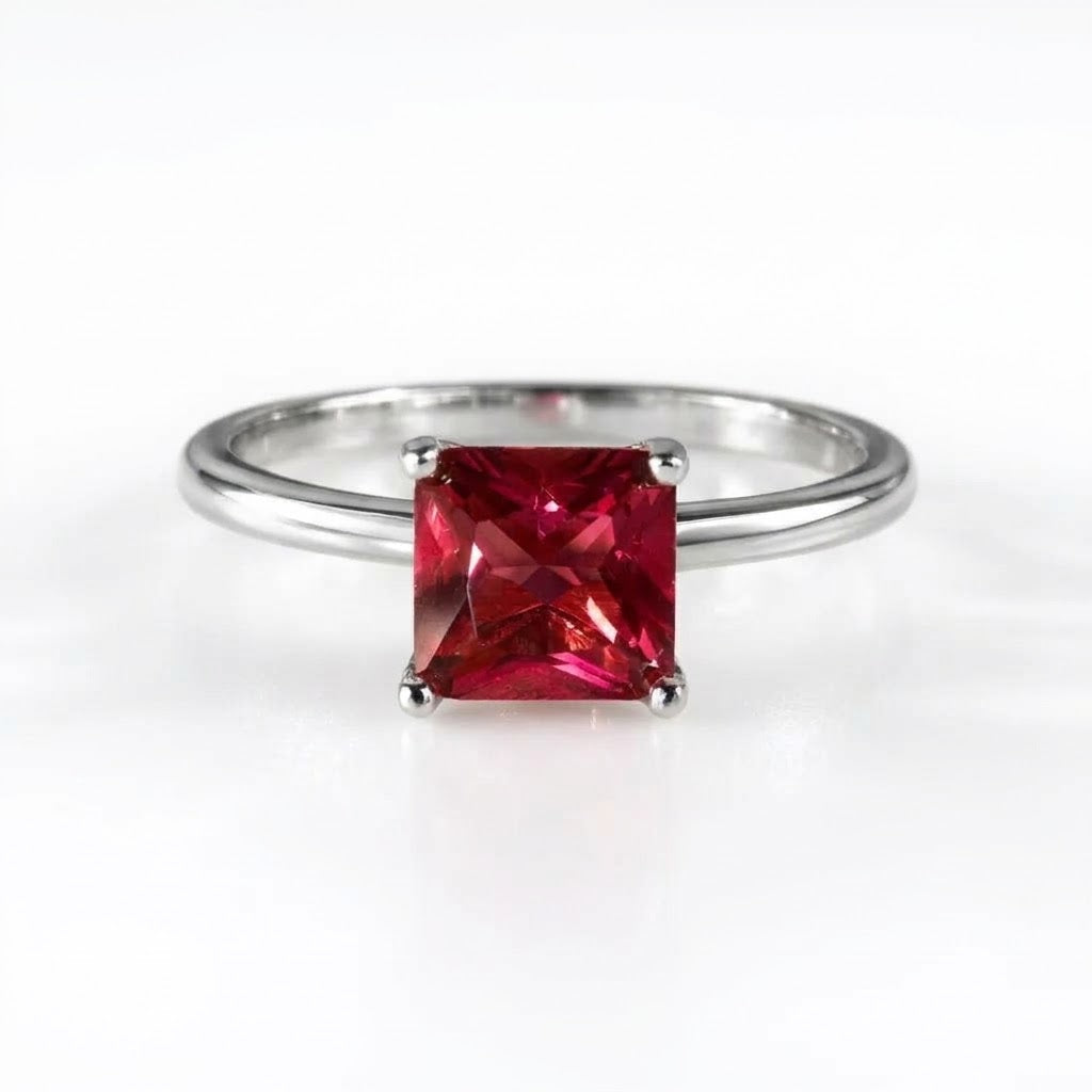 A silver band ring featuring a square-cut, vibrant red gemstone in a four-prong setting, photographed on a clean white background.