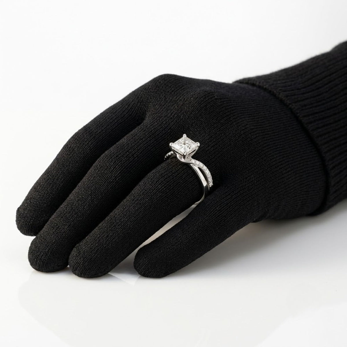 Princess cut diamond solitaire engagement ring set in white gold or platinum with a twisted, bypass pavé diamond band, displayed on a black-gloved hand.