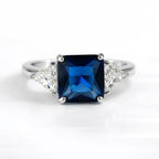 A silver ring featuring a square-cut deep blue sapphire center stone flanked by two triangular clear diamonds on a white background.