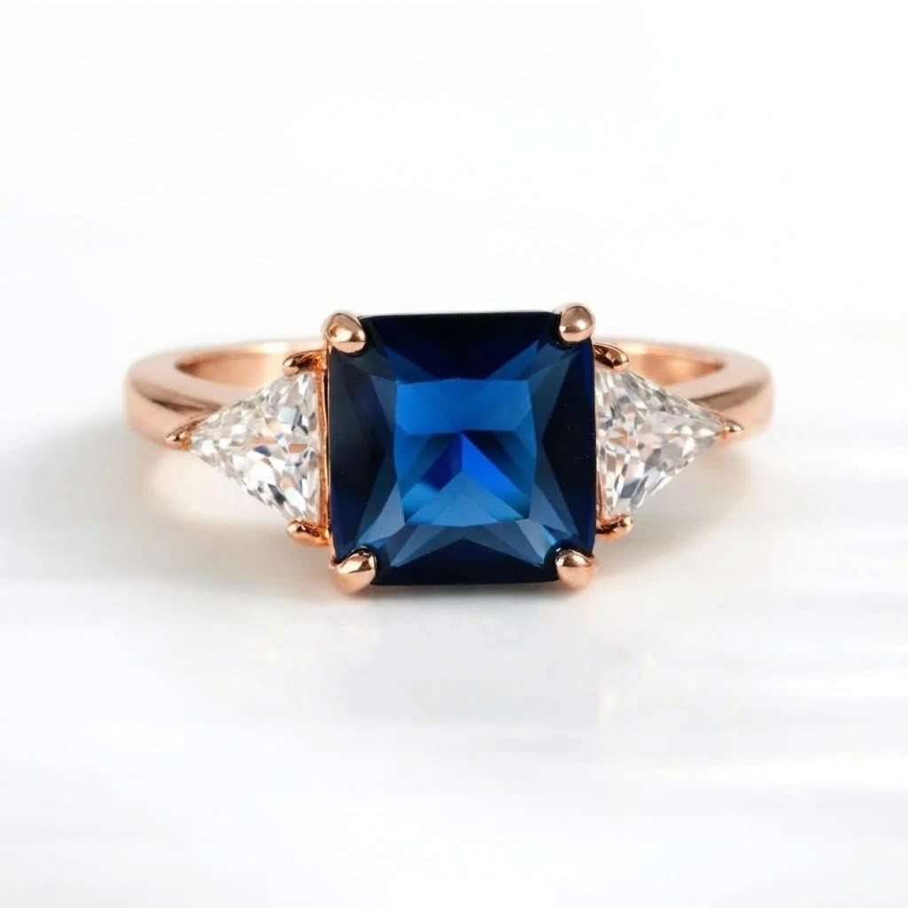 A rose gold ring featuring a square-cut deep blue sapphire center stone and two clear triangular side stones, set against a clean white background.