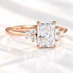 Angled view of a radiant cut diamond center stone in a four-prong rose gold setting with a cluster of three round side stones on each side