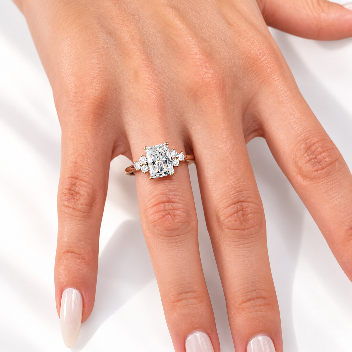Radiant cut diamond center stone in a four-prong rose gold setting, flanked by a cluster of round side stones on each side