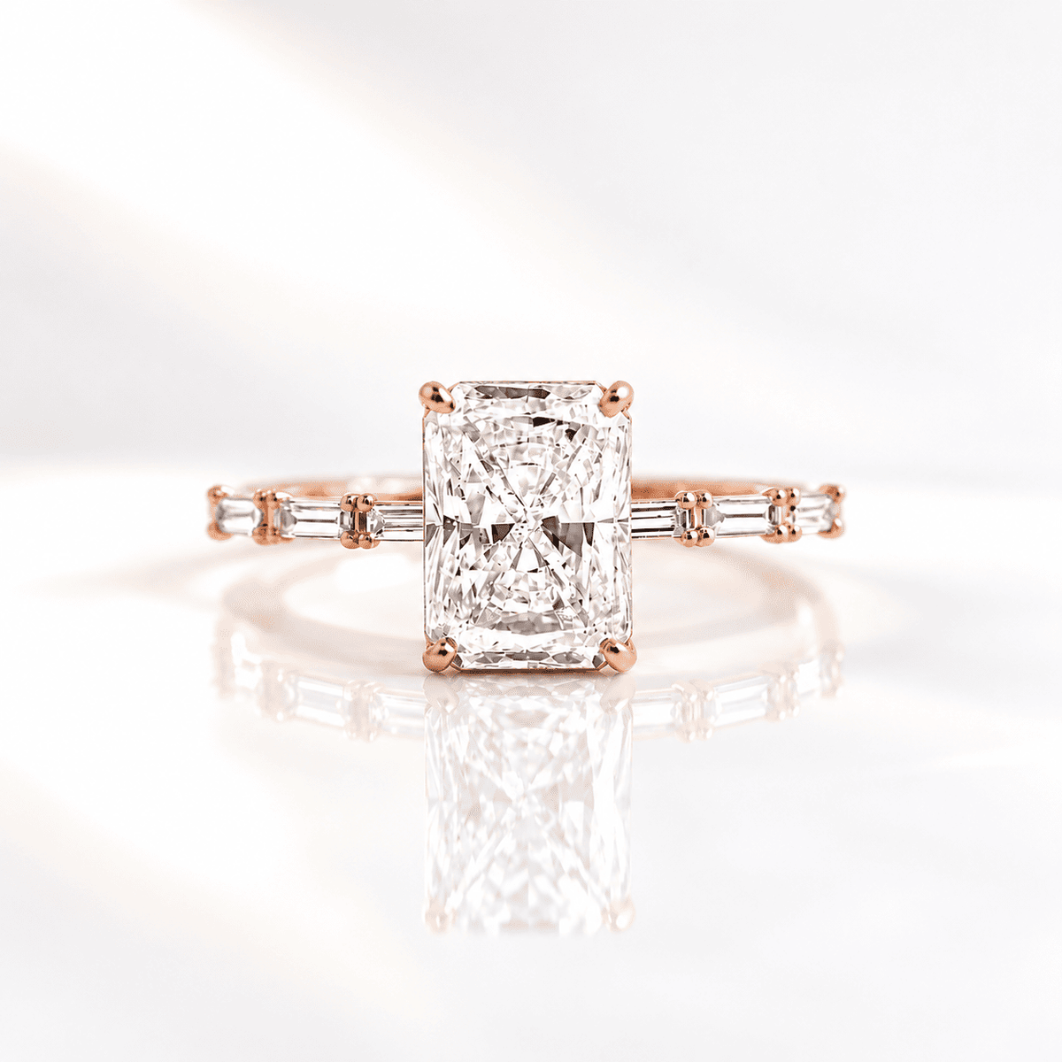 Radiant cut diamond engagement ring with four prongs and baguette side stones on a slim band, set in rose gold on a soft, reflective white background.
