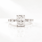 Radiant cut diamond engagement ring with four prongs and baguette side stones on a slim band, set in white gold on a reflective white surface.S