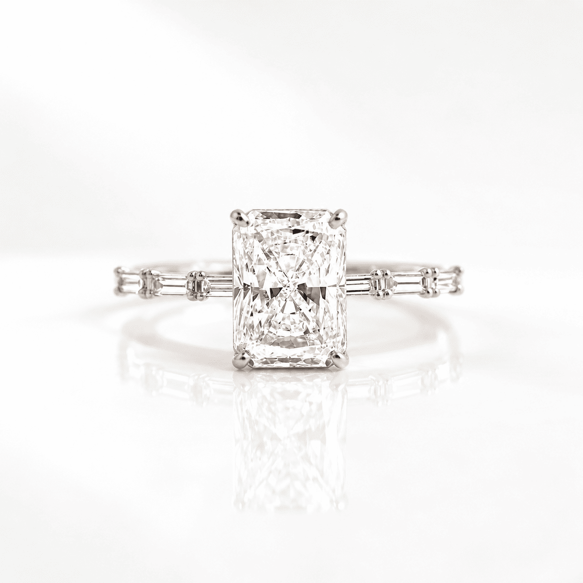 Radiant cut diamond engagement ring with four prongs and baguette side stones on a slim band, set in white gold on a reflective white surface.S