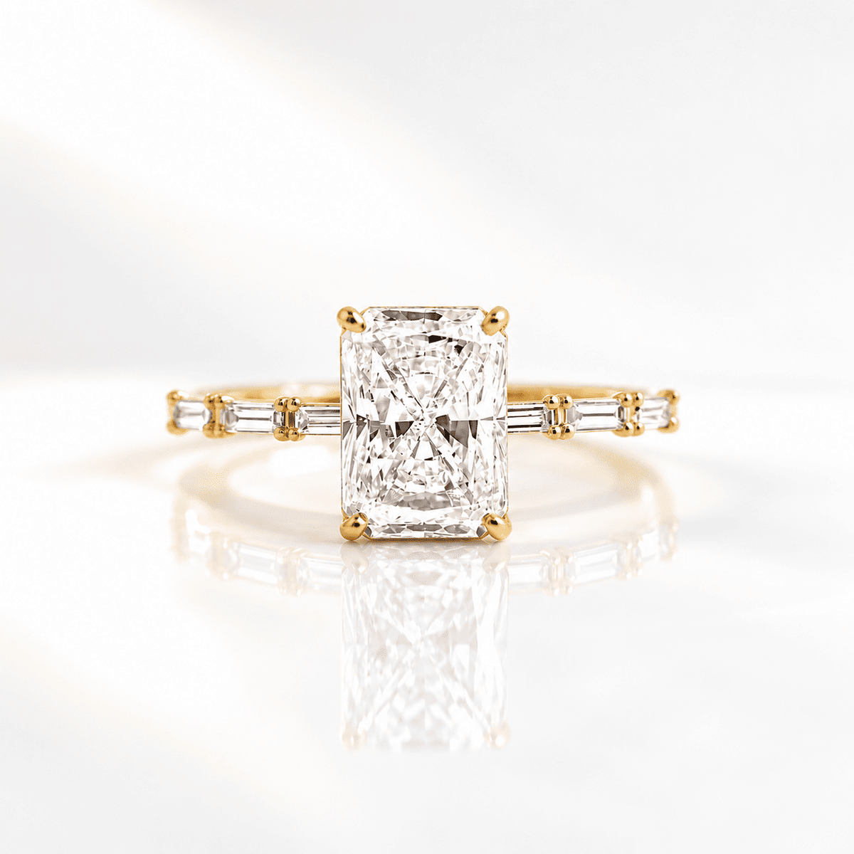 Radiant cut diamond engagement ring with four prongs and baguette side stones on a slim band, set in yellow gold on a glossy white surface.