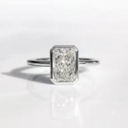 An emerald-cut diamond ring in a silver-toned setting on a clean white reflective surface.